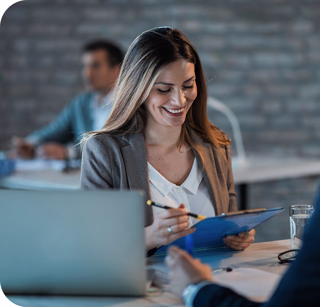 Professional woman sitting at a desk, smiling while looking down at her clipboard.